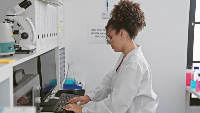 A young woman scientist in a lab coat works on a computer in a modern laboratory setting, surrounded by scientific equipment.