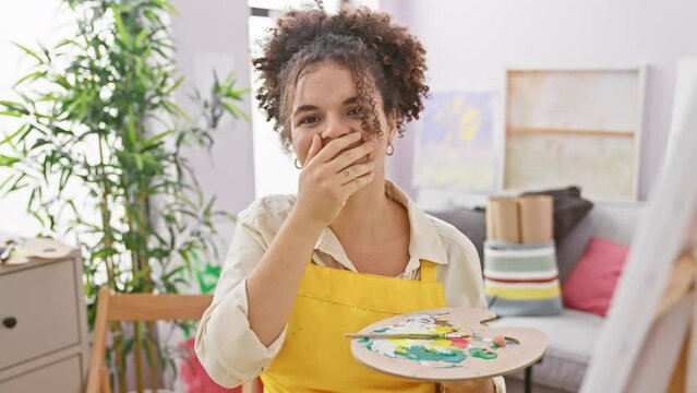 Laughing artist with curly hair, covering mouth in a fun embarrassed gesture at studio. hispanic woman sitting joyfully amidst gossip