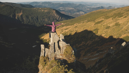 Aerial tourist woman rise up hands. Girl sit on stone at mountain top. Nature landscape. Mount rock peak. Traveler mountaineering, climbing. Tourism and travel lifestyle. Carpathians, Ukraine, Europe - obrazy, fototapety, plakaty