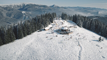 Winter active sport resort at snow mountain top aerial. People at nature landscape. Ski slope at fir forest. Mount ranges with pine trees. Tourist attraction at Carpathians, Bukovel, Ukraine, Europe