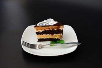 A piece of chocolate biscuit cake with mint leaves and a fork on a white ceramic plate on a black background.