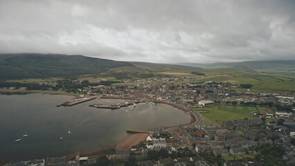 Port citycsape with modern, historic buildings at ocean bay aerial. Ships, yachts at wharf. Streets with old buildings at road with cars, trucks. Green hill, valley at Campbeltown pier city, Scotland