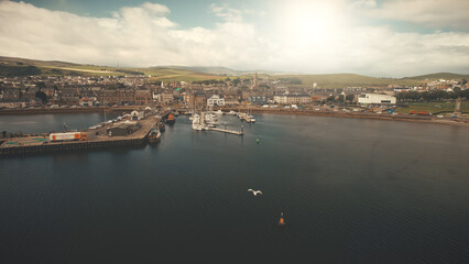 Sun pier town cityscape aerial. Seagull flight over ocean bay. Yachts, ships at marina. Historic buildings architecture landmark at urban streets of Campbeltown city, Scotland. Cinematic seascape