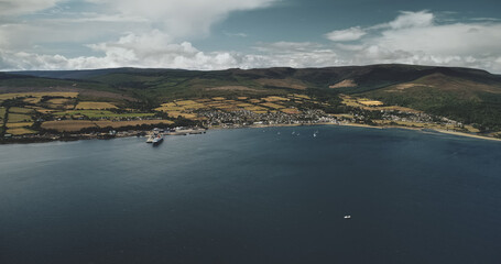 Brodick ocean ferry pier aerial view: ships, boats and sailboat on ocean surface at harbor. Wonderful Scottish scape of nature with forests and valleys in summer shot