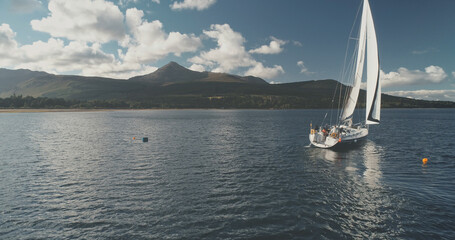 Yacht racing at ocean bay coast aerial. Passengers on sailboat at open sea at summer cloudy day. Brodick harbor at mountain island of Arran, Scotland. Cinematic scenery of luxury cruise on sail boat.