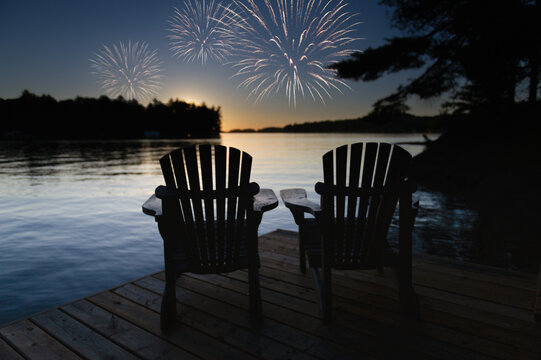 Canada Day celebration fireworks light up the sky over Muskoka Lake, Ontario, as two Adirondack chairs on the dock offer a front-row seat to the spectacular show.