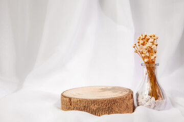 Transparent vase with stones inside and small white flowers with yellow handles and raw wood on the side with a white fabric background. Space for writing or product.