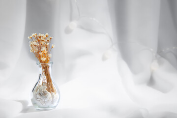 Transparent vase with stones inside and small white flowers with yellow handles on a white fabric background and blurred lights. Space for writing.