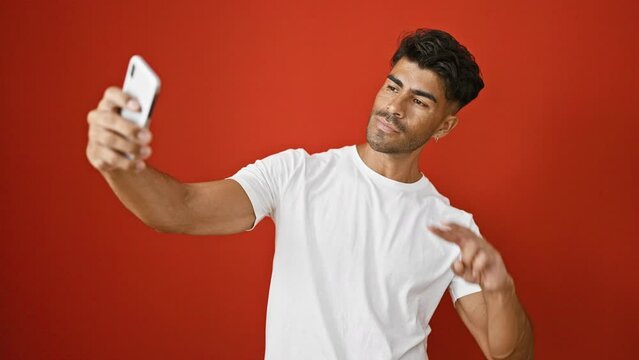 Handsome young latino man with beard in white shirt taking selfie against red background