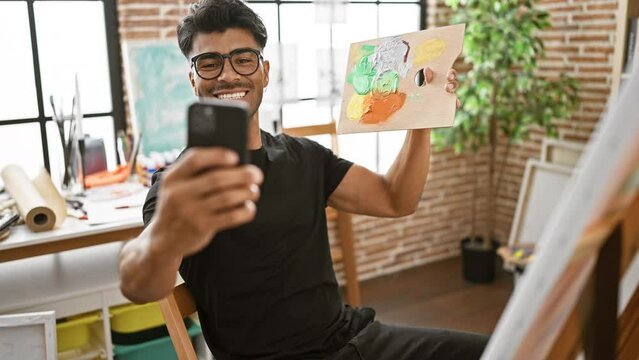 Handsome latin man with beard and glasses takes selfies in an art studio, holding a paint palette while smiling.