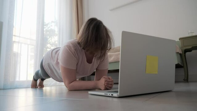 Focused young woman doing plank exercise at home in front of her laptop, maintaining fitness while indoors.