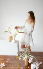 young elegant woman in white dress with spring flowers in vases in white room - 782720031