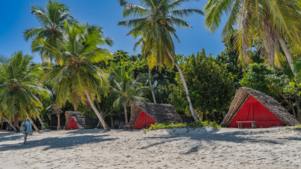Tourist bungalows with triangular thatched roofs and cloth curtains stand in a row. A man walks along a sandy beach with a backpack and flip-flops in his hand. Palms, tropical vegetation, blue sky