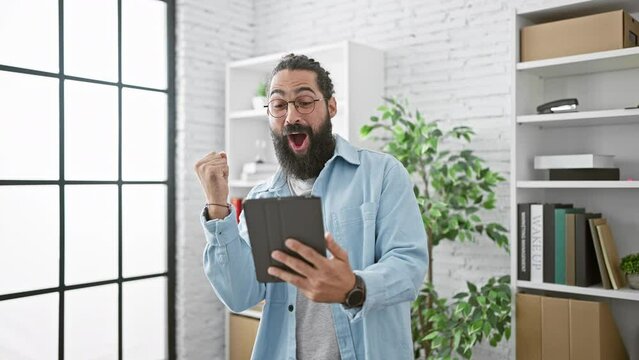 Hispanic man with beard and glasses in a light blue shirt showing different emotions while using a tablet in a modern office.