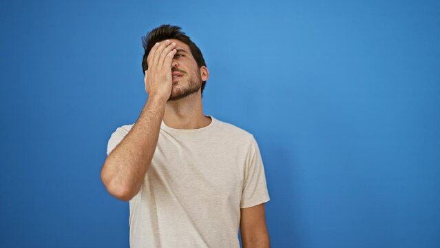 A stressed hispanic adult man with a beard in a casual t-shirt against a blue wall outdoors expressing exhaustion.