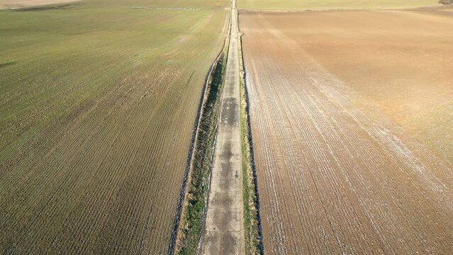 Hovering above the landscape, this drone footage presents a serene aerial view of a solitary country road cutting a straight path through the contrasting textures of winter fields. The muted colors of