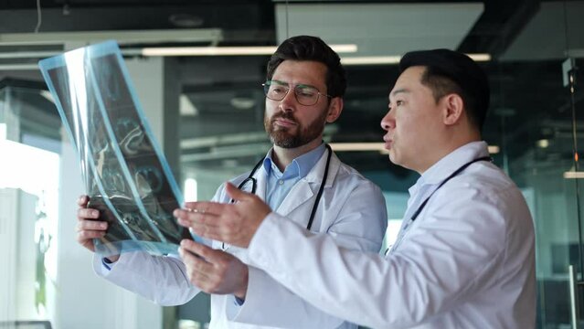 Side view portrait of gentle caucasian and asian males doctors examining x-ray in specially equipped room. Determinative men discussing problem of difficult patient indoors. Concept of diagnosis.