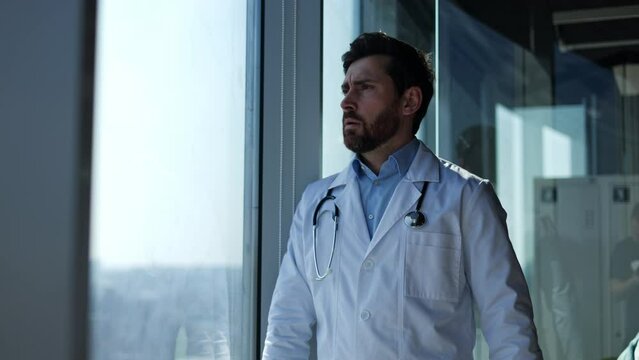 Handsome man in white coat finishing reading medical papers and taking off glasses while standing by window. Frustrated family doctor thinking about new strategy for curing difficult patient.