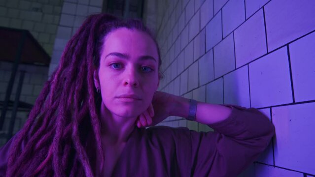 Zoom-in portrait of young dreadlocked Caucasian woman in coverall leaning on tiled wall and posing for camera in dark room with neon light