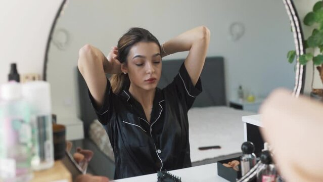 portrait of a young beautiful woman in pajamas sitting in front of the mirror, styling her luxurious hair into a bun, pinning it up, and posing gracefully