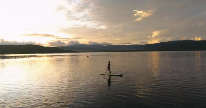 Woman Standing On Paddleboard At Daybreak On Lake With Silhouette Of Kayaker In Distance. Moso Island, Vanuatu. drone pullback, tilt-up shot