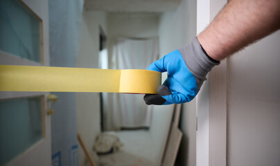 Worker with a masking tape in hands at construction site. Quick and easy painting process. Accessories for install furniture, repair home. Man dressed in blue gloves. Home renovation concept