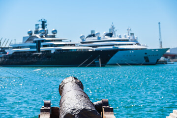 Eine alte Kanone im Hafen, im Hintergrund liegen Luxusyachten vor Anker, Tarragona, Spanien - obrazy, fototapety, plakaty