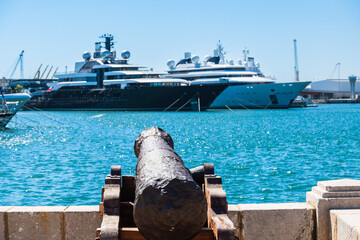 Eine alte Kanone im Hafen, im Hintergrund liegen Luxusyachten vor Anker, Tarragona, Spanien - obrazy, fototapety, plakaty