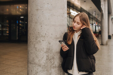 Half length portrait of girl in casual wear holding smartphone for blogging in the city in the autumn-winter season. Teen surfing the internet outdoors - 782856467