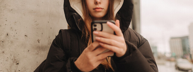 Half length portrait of girl in casual wear holding smartphone for blogging in the city in the autumn-winter season. Teen surfing the internet outdoors
