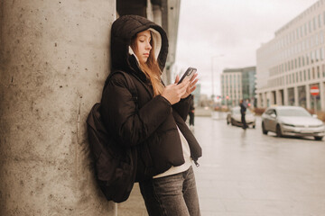 Half length portrait of girl in casual wear holding smartphone for blogging in the city in the autumn-winter season. Teen surfing the internet outdoors - 782856885