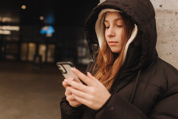 Half length portrait of girl in casual wear holding smartphone for blogging in the city in the autumn-winter season. Teen surfing the internet outdoors - 782857025
