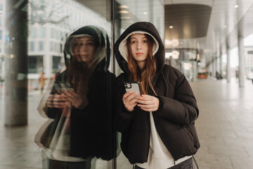 Half length portrait of girl in casual wear holding smartphone for blogging in the city in the autumn-winter season. Teen surfing the internet outdoors - 782857210