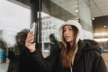 Half length portrait of girl in casual wear holding smartphone for blogging in the city in the autumn-winter season. Teen surfing the internet outdoors - 782857402