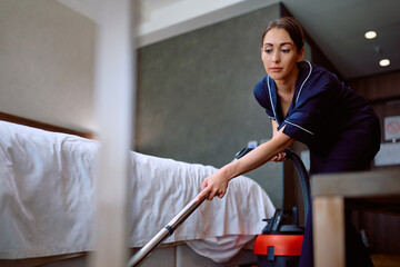 Young chambermaid vacuuming hotel room.