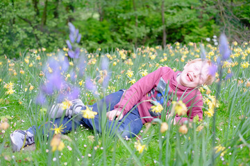 A young boy with fair hair and a wide grin lies on his back among bright yellow daffodils in a verdant park. He is casually dressed in a maroon hoodie and jeans, his pose relaxed and joyful amidst the