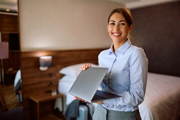 Happy businesswoman using laptop in hotel room and looking at camera.