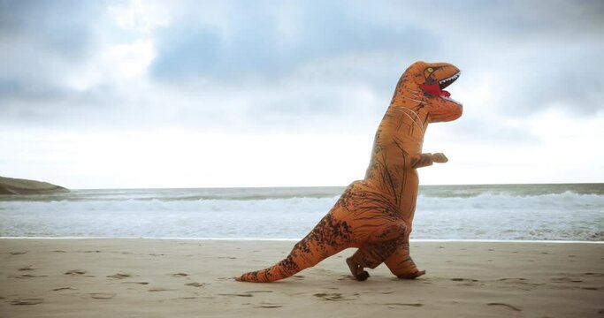 Dinosaur costume, beach and funny with sunshine, comic and running with waves and sand. Blue sky, Halloween outfit and humor with seaside and travel with vacation and nature with comedy and holiday