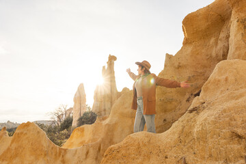 A woman stands on a rocky hillside, wearing a brown jacket and jeans. She is smiling and pointing to the sky, which is filled with sunlight. Concept of happiness and appreciation for the beautiful day