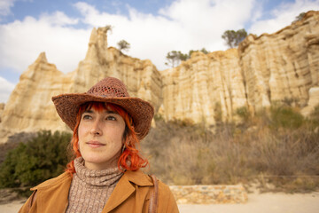 A woman with red hair and a cowboy hat stands in front of a mountain. She is wearing a brown coat...