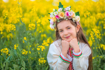 Dreaming beautiful young girl with spring flower chaplet, ethnic folklore dress with traditional bulgarian embroidery on a yellow rapeseed field - 782872856