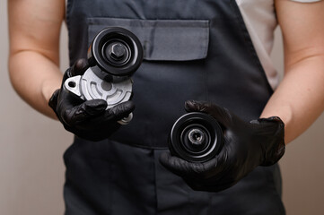 An auto mechanic holds in his hands in black gloves a idler pulley with a tensioner for a car's poly V-belt and a parasitic roller, close-up. Auto parts replacement concept, car maintenance. - obrazy, fototapety, plakaty