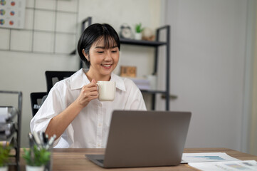 An adult woman sips her coffee while engaging with a laptop in a bright home office, reflecting a...