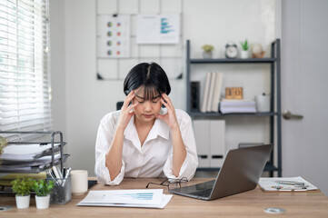 Exhausted female office worker feeling stressed, holding glasses, with laptop and documents on desk.