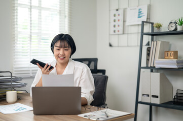 Cheerful businesswoman in white shirt using smartphone with laptop and documents on her office desk.