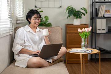 An adult woman sips her coffee while engaging with a laptop in a bright home office, reflecting a...