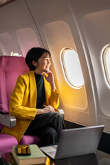 Smiling businesswoman in a yellow blazer using a laptop on a tray table, working efficiently on an airplane.