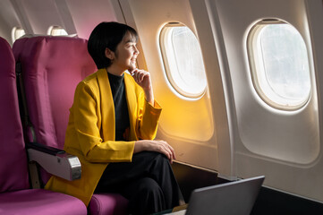 Smiling businesswoman in a yellow blazer using a laptop on a tray table, working efficiently on an...