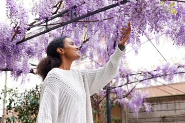 A cherry black afro woman is holding a blooming purple wisteria flower hanging in the metal garden...