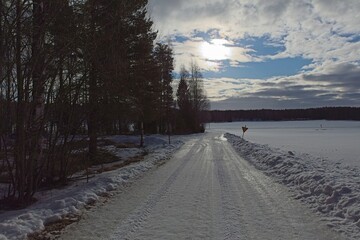 Plowed rural road with tire tracks that is lined with trees in winter with clouds in the sky and...